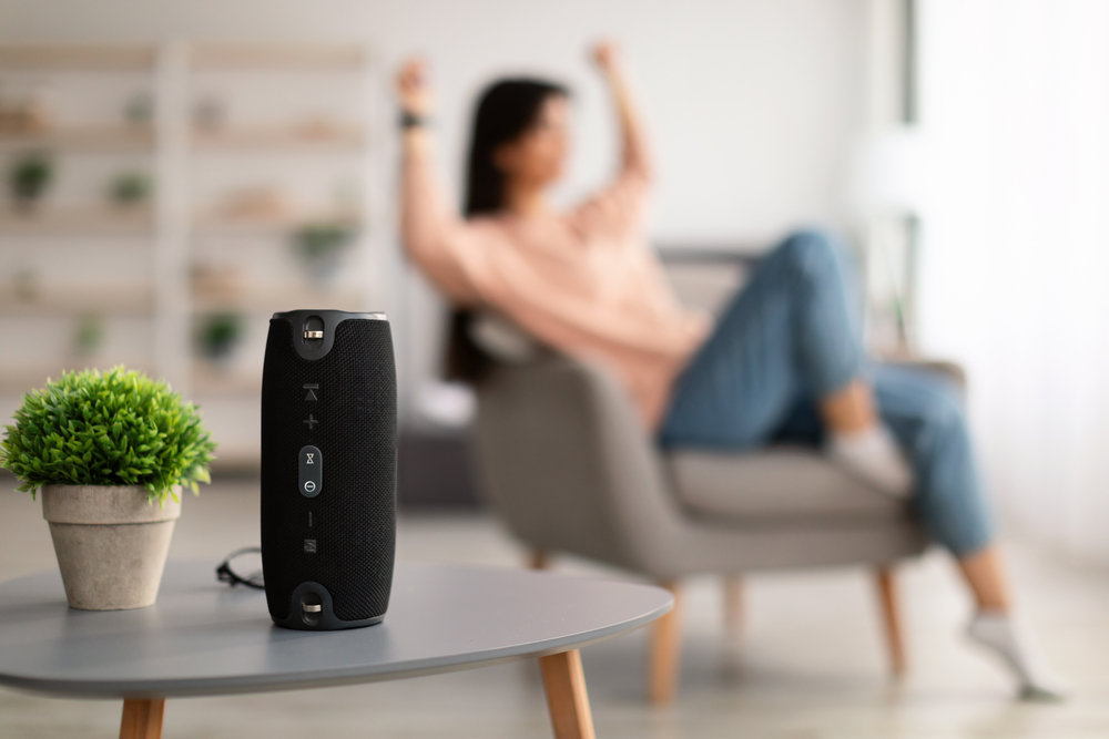 Closeup of smart portable wireless speaker on the table, selective focus. Young woman listening to music and dancing, sitting on armchair in the blurred background